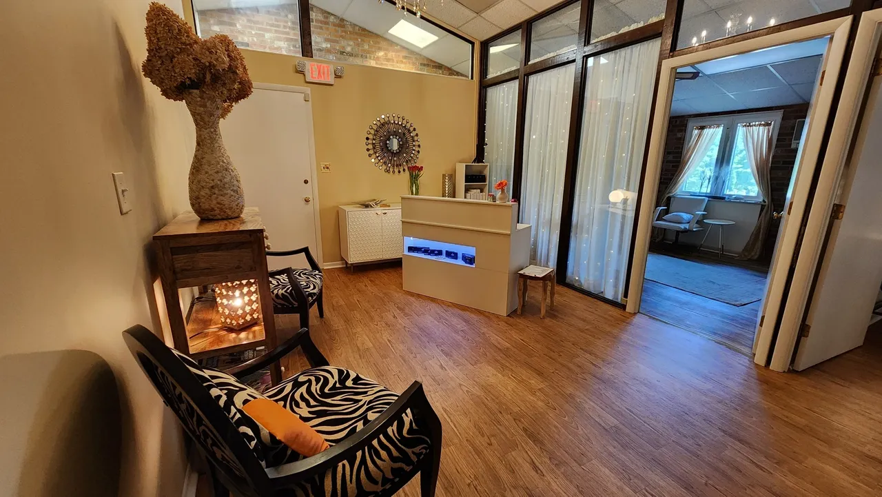 Reception area with a small desk, chairs with zebra print cushions, and a vase filled with dried flowers. A wall mirror adds depth, reflecting the serene view of an adjacent room. Neutral tones and wooden flooring enhance the cozy atmosphere, perfect for offering luxury skincare services.