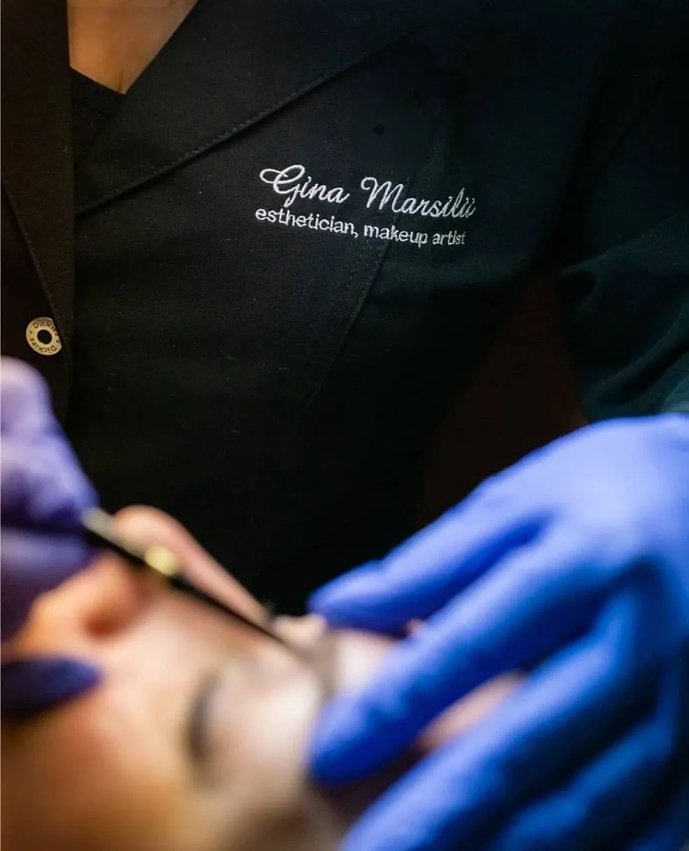 Close-up of an esthetician with "Gina Marsili, esthetician, makeup artist" embroidered on their jacket, applying a luxury skincare treatment to a client's face while wearing blue gloves.