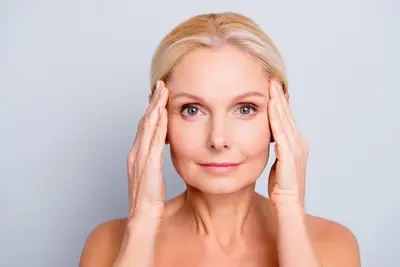 A woman with blonde hair, radiant and flawless from luxury skincare, holds both hands to the sides of her face while looking forward against a plain background.