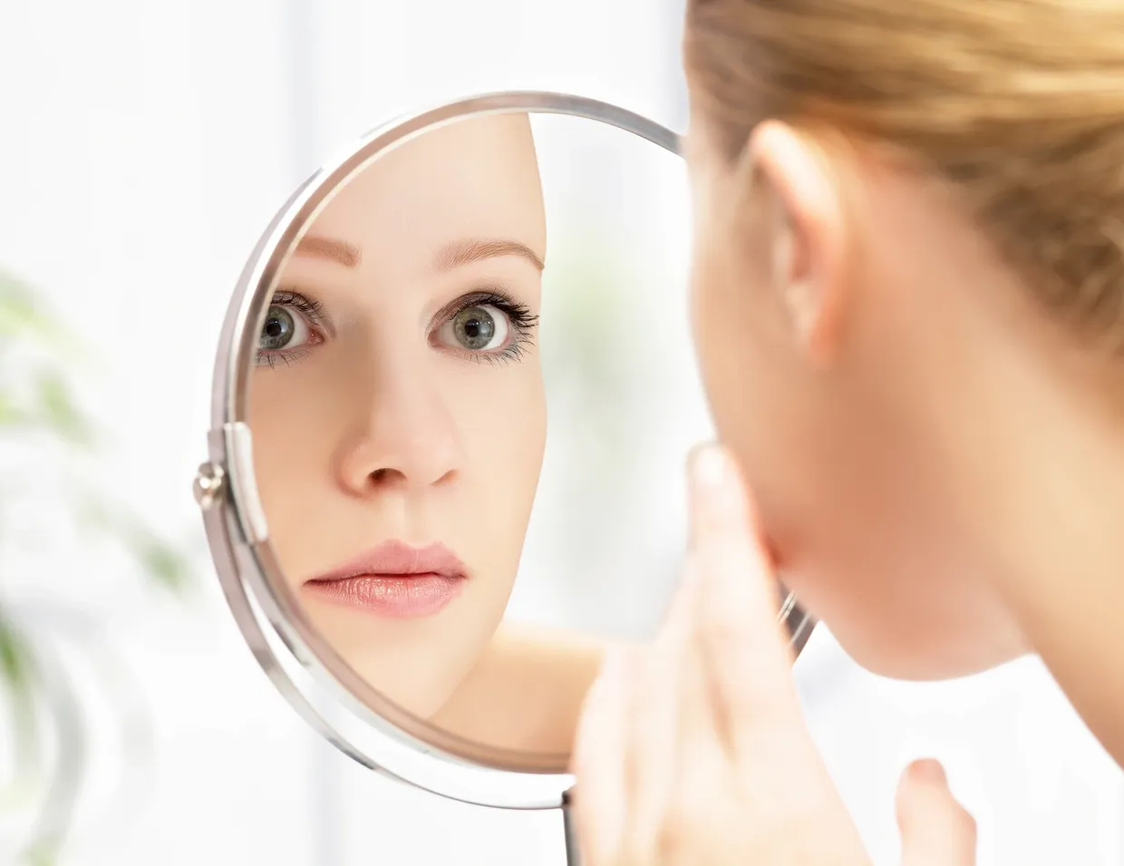 A person looks at their face in a round mirror, touching their chin with a neutral expression, contemplating the results of luxury skincare.