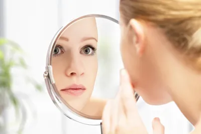 A person looks at their reflection in a round mirror, focusing on their face with a slightly concerned expression. The background is blurred, hinting at the elegance of their luxury skincare products subtly arranged behind them.