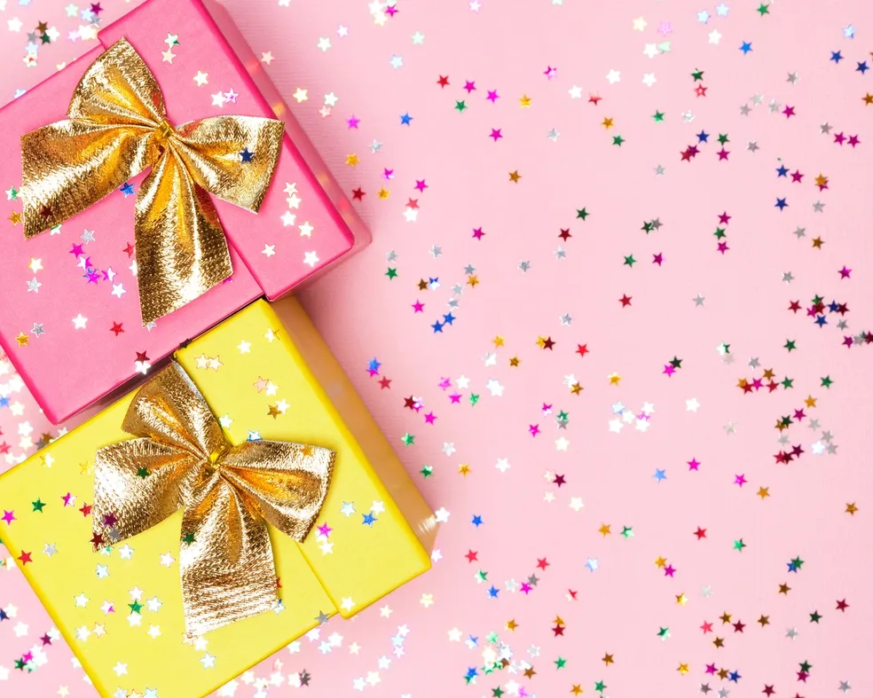 Two gift boxes with gold bows, one pink and one yellow, on a pink background with scattered multicolored star-shaped confetti. Perfectly presented for luxury skincare gifts.