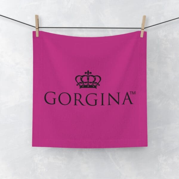 A Face Towel in pink, featuring a crown logo and the text "GORGINA™," is hanging on a clothesline against a light background.