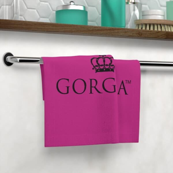 A pink Face Towel embossed with the word "GORGA™" and a crown logo, displayed on a rack against a tiled background with a shelf of toiletries.