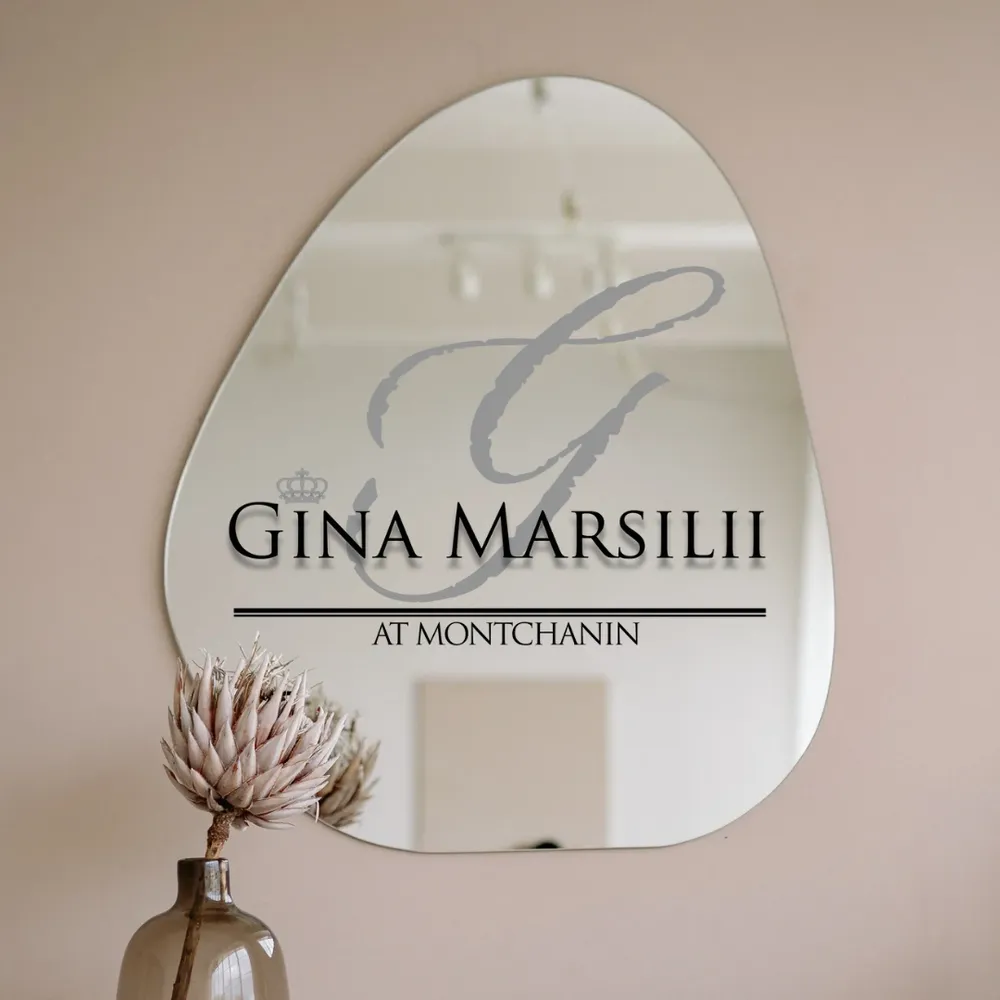 Irregularly shaped mirror with "Gina Marsilii at Montchanin" text, reflecting ceiling lights. A dried flower in a vase sits elegantly in the foreground, evoking the serene ambiance of luxury skincare rituals.